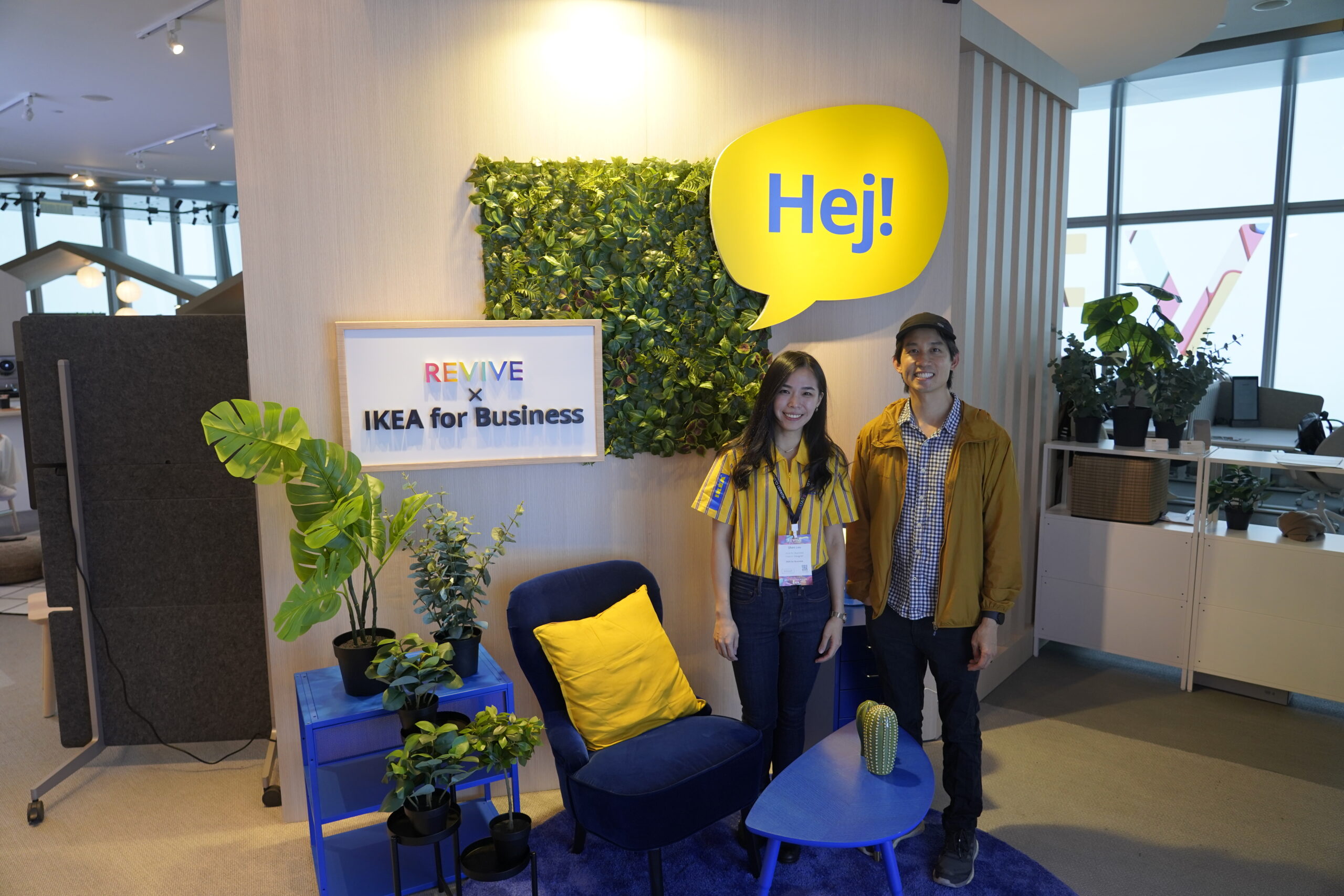 Two individuals standing in front of a promotional display for REViVE IKEA for Business, featuring a living wall, furniture, and a large speech bubble with the word "Hej!"





