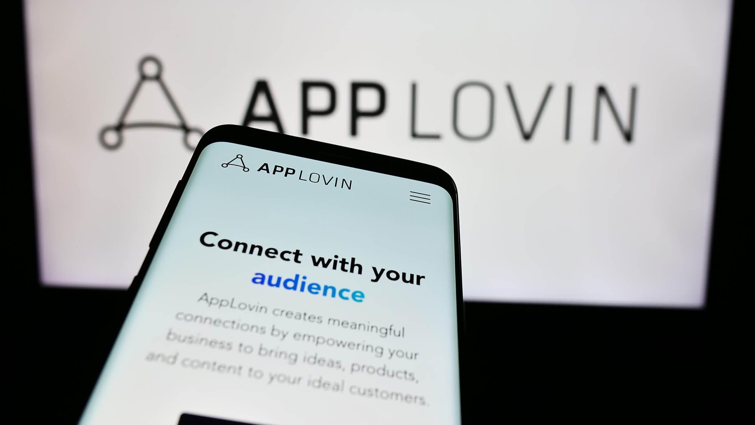 Mobile phone with webpage of US mobile technology company AppLovin Corporation in front of business logo. Focus on top-left of phone display.