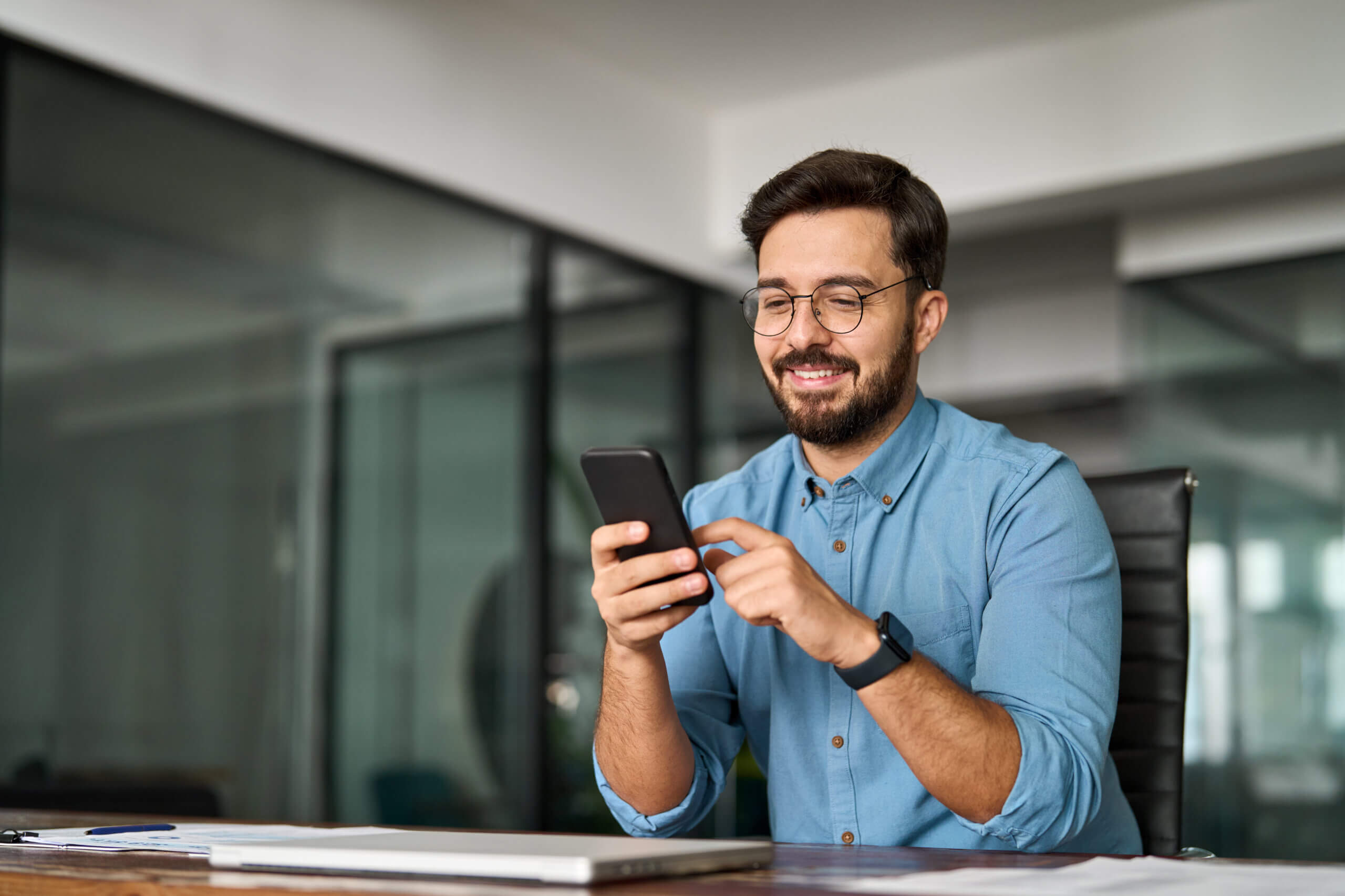 Busy young Latin business man holding smartphone working in office. Professional happy businessman entrepreneur or manager using financial banking apps making easy payments on cell phone at work.
