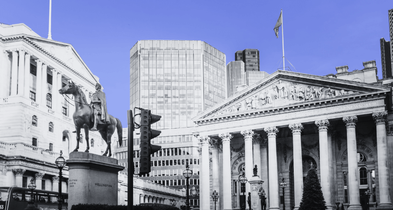 The Royal Exchange in London with its grand columns and triangular pediment, alongside the equestrian statue of the Duke of Wellington, shown in black and white with a blue sky overhead.