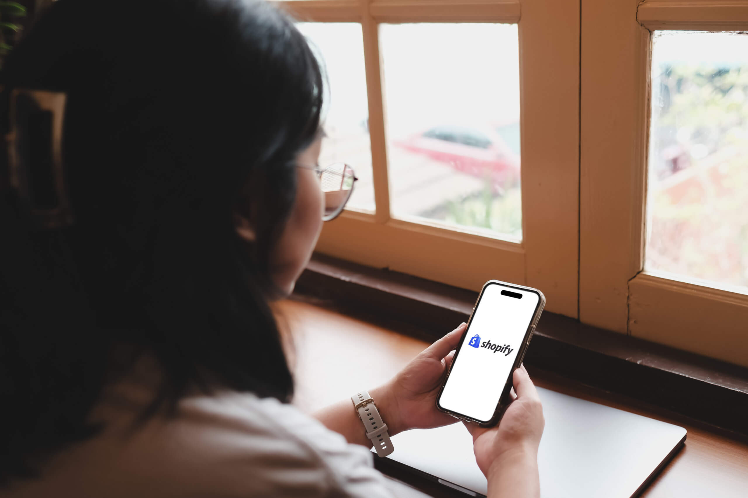 Bangkok, Thailand. April 4, 2025; Woman holding smartphone showing Shopify logo while sitting by the window with closed laptop on table in cozy cafe.