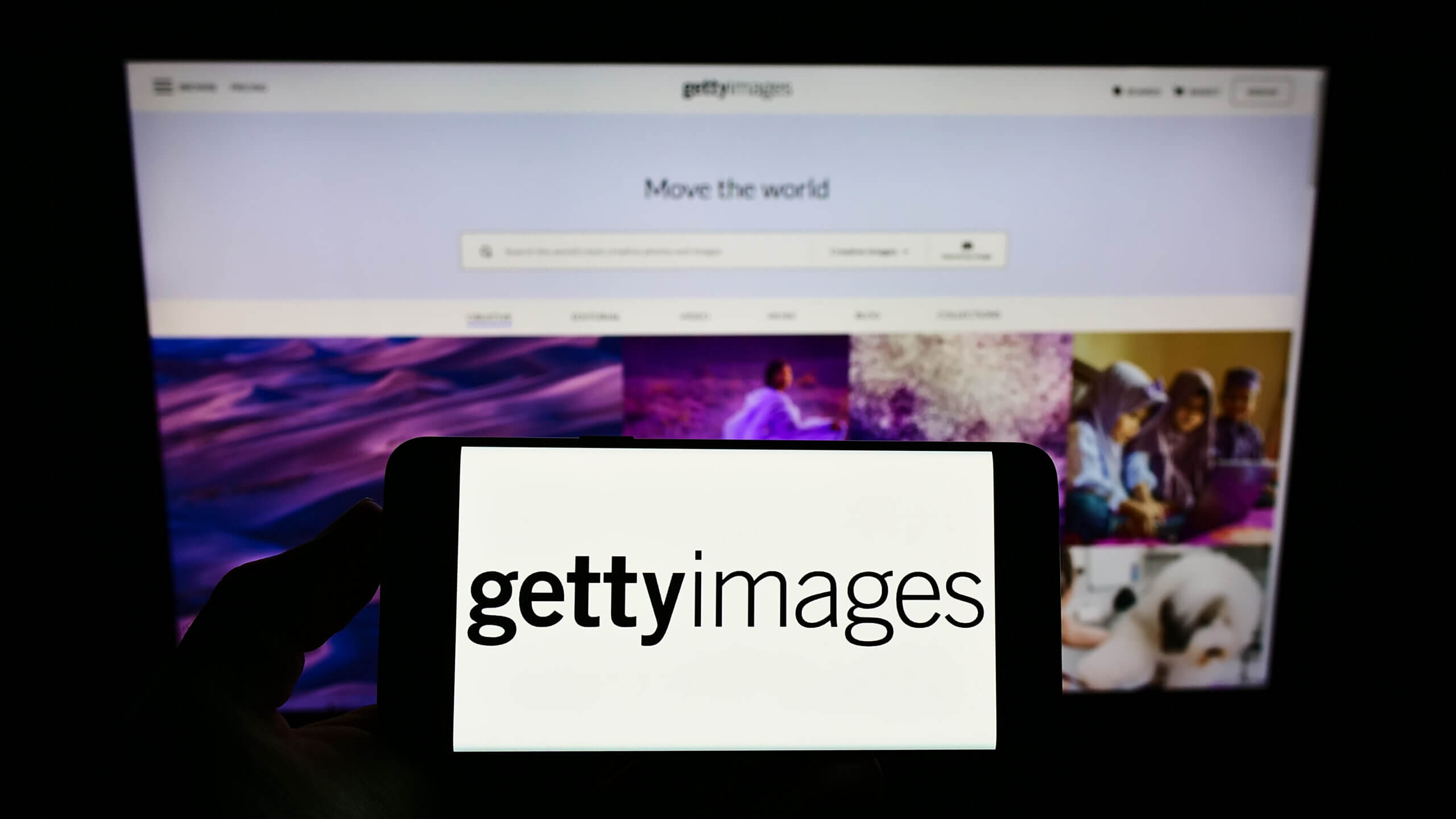 A person holds a smartphone displaying the Getty Images logo in front of a blurred computer screen showing the Getty Images website homepage with the slogan “Move the world.”