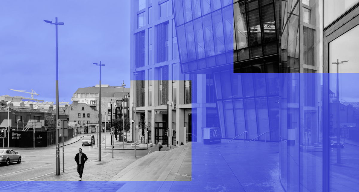 Black and white city street scene with a lone pedestrian walking past tall modern glass buildings, overlaid with translucent blue geometric blocks.