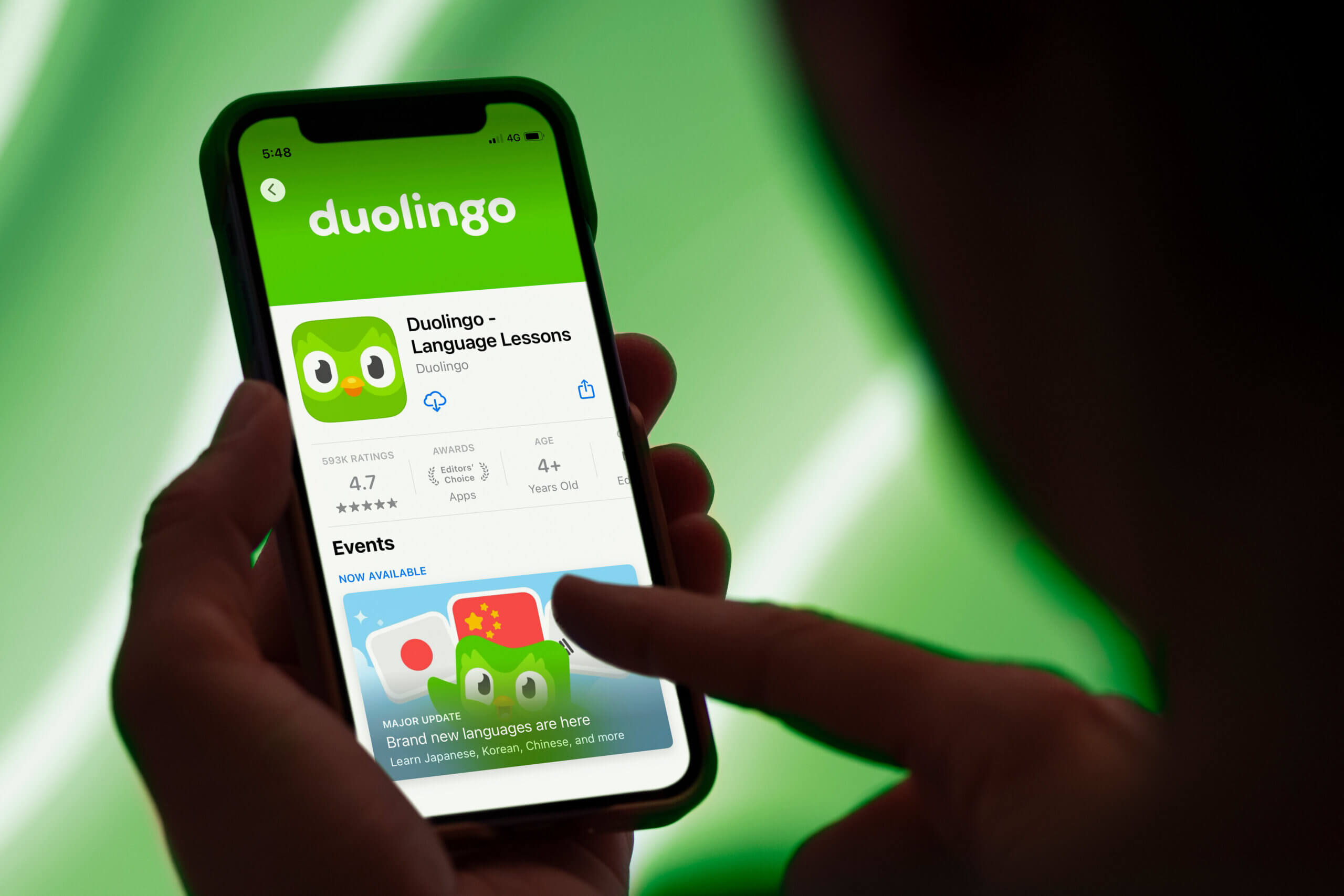 A person is holding a smartphone displaying the Duolingo app page in the App Store. The screen shows the Duolingo owl mascot, the app name "Duolingo – Language Lessons," a 4.7-star rating from nearly 600,000 reviews, and an Editors’ Choice award. Below, an event promotion highlights a major update introducing new languages with illustrated flags of Japan and China alongside the green Duolingo owl. The background is brightly lit in green, matching the app’s branding.