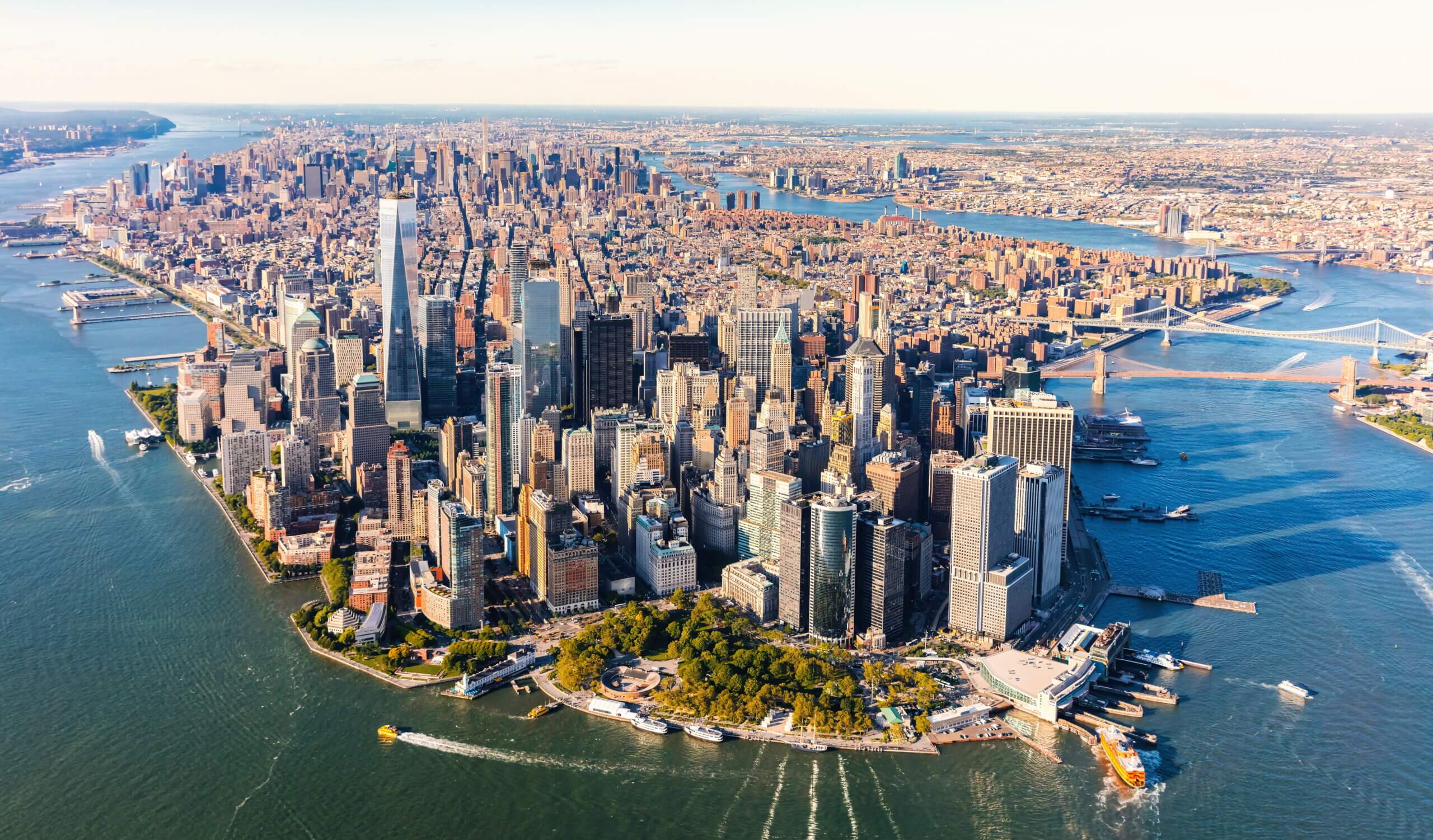 Aerial view of Lower Manhattan in New York City on a clear day, showing dense clusters of skyscrapers surrounded by rivers. The tip of Manhattan features Battery Park with green trees, ferries, and piers along the waterfront. The Brooklyn and Manhattan Bridges are visible spanning the East River in the background, while the Hudson River extends along the left side with boats leaving white trails in the water.