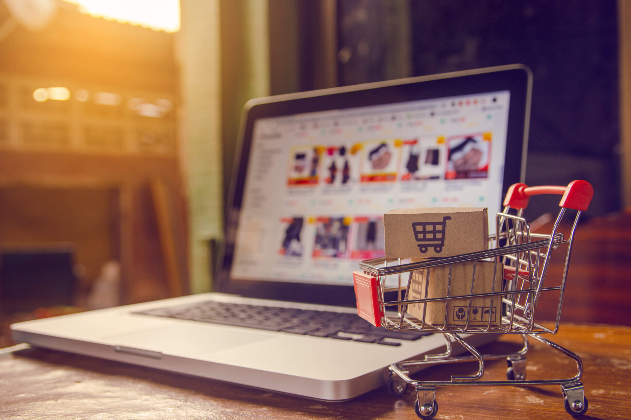 A miniature shopping cart filled with small cardboard boxes sits on a wooden table in front of an open laptop displaying an online shopping website. The scene is warmly lit by sunlight coming through a window in the background.