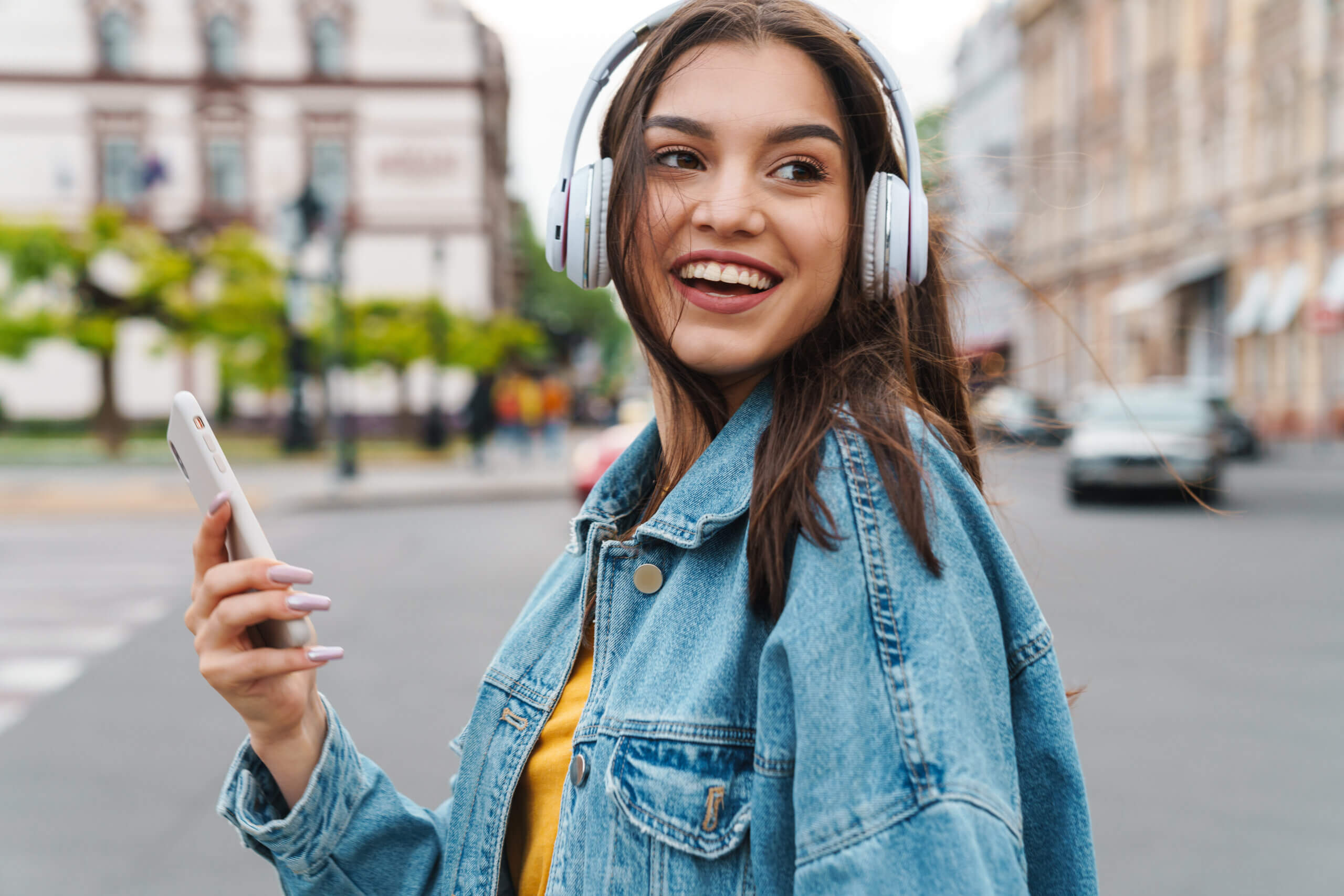 Image of nice laughing woman listening music with smartphone and wireless headphones on city street