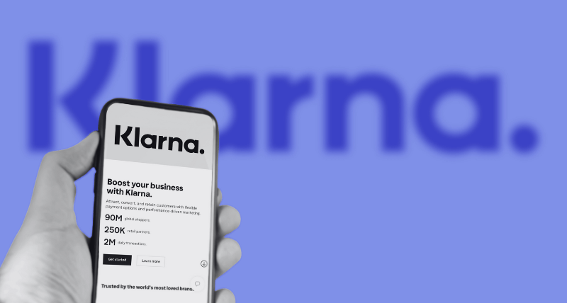 A hand holding a smartphone displaying Klarna’s business page, with the Klarna logo enlarged and blurred in the blue background.