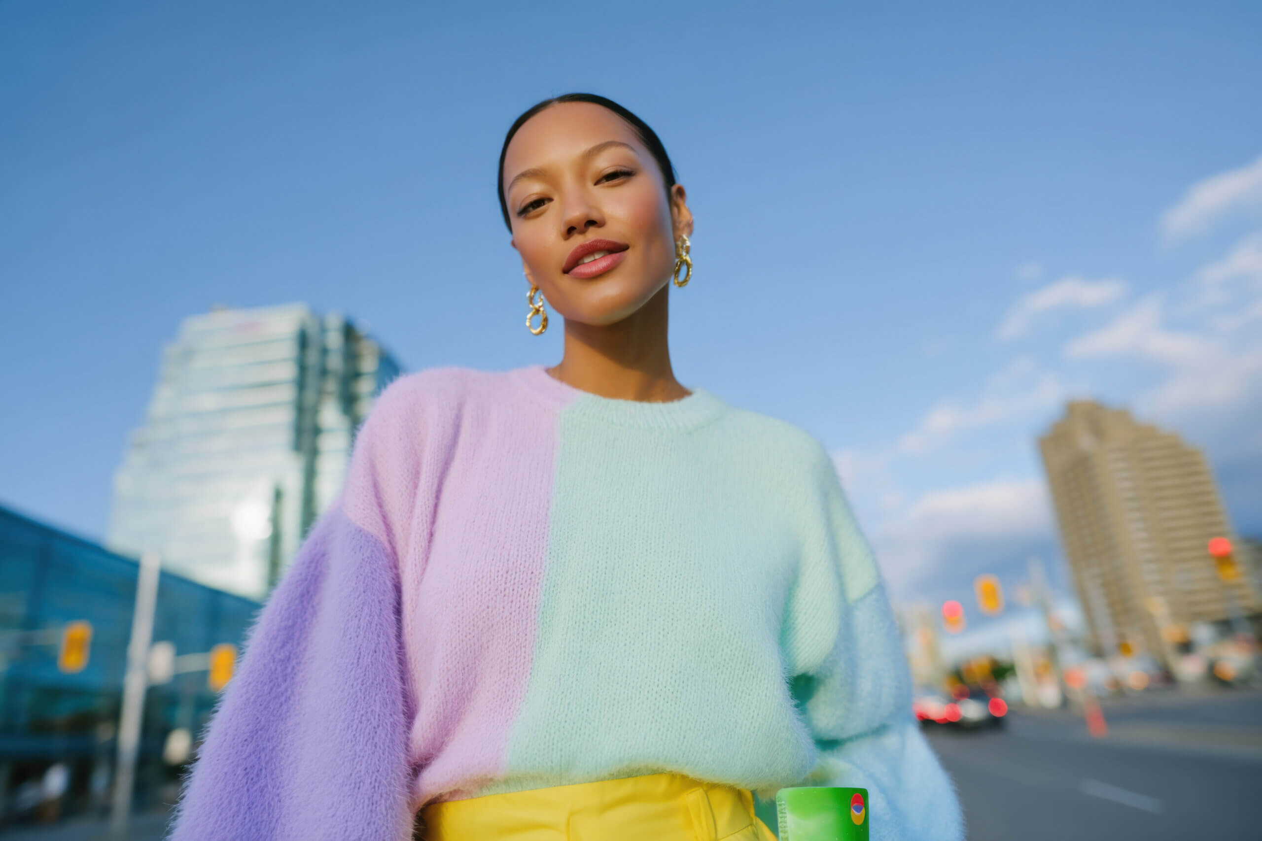 Young woman showcasing a vibrant pastel sweater paired with yellow pants, posing confidently against an urban city backdrop
