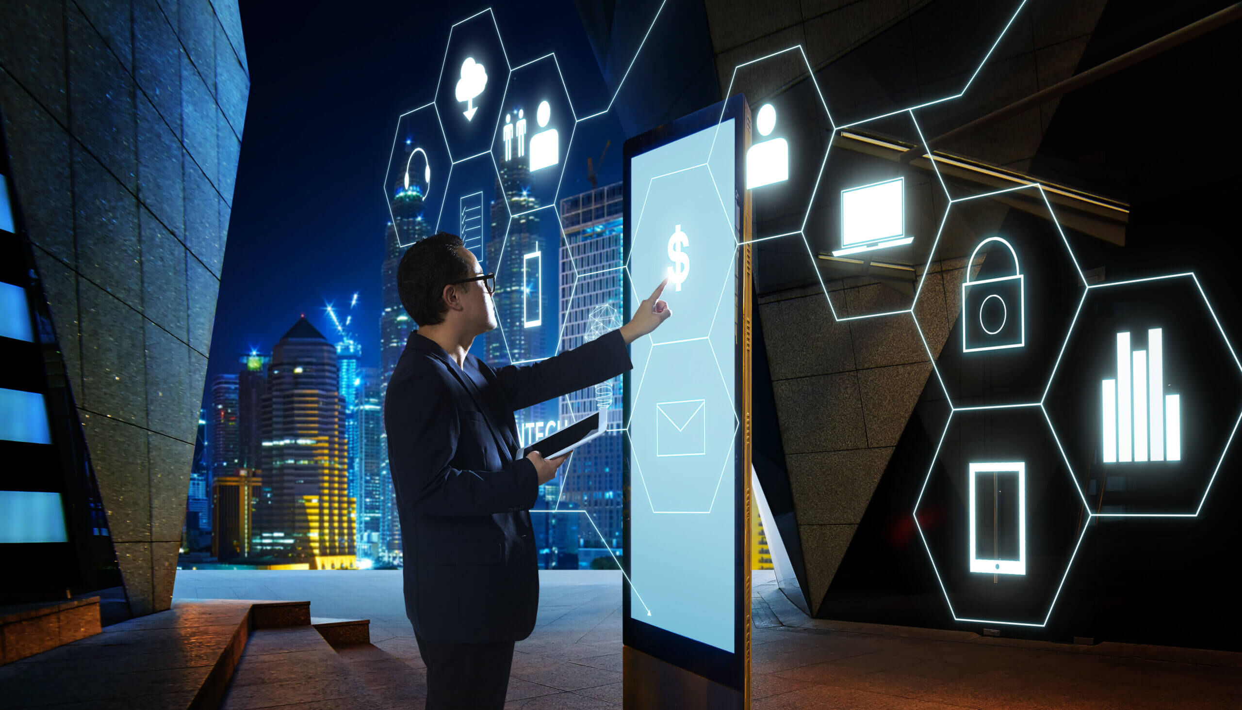A man in a dark suit interacts with a large vertical touchscreen displaying a dollar sign, standing in a modern outdoor cityscape at night. He holds a tablet in one hand and reaches toward the screen with the other. Around him, glowing hexagonal digital icons—such as a lock, cloud, computer, bar chart, and user profile—float in the air, suggesting futuristic financial or technology interfaces against the backdrop of illuminated skyscrapers.