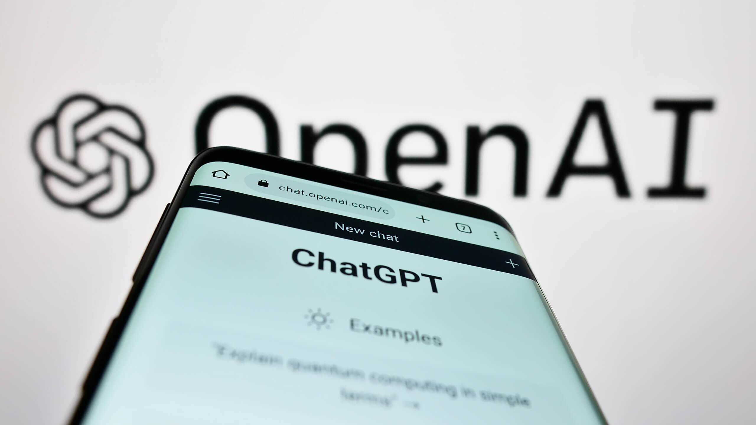 A smartphone displaying the ChatGPT interface is held up close to the camera, with the OpenAI logo blurred in the background. The phone screen shows the ChatGPT homepage with navigation icons and the text “New chat” and “ChatGPT.”