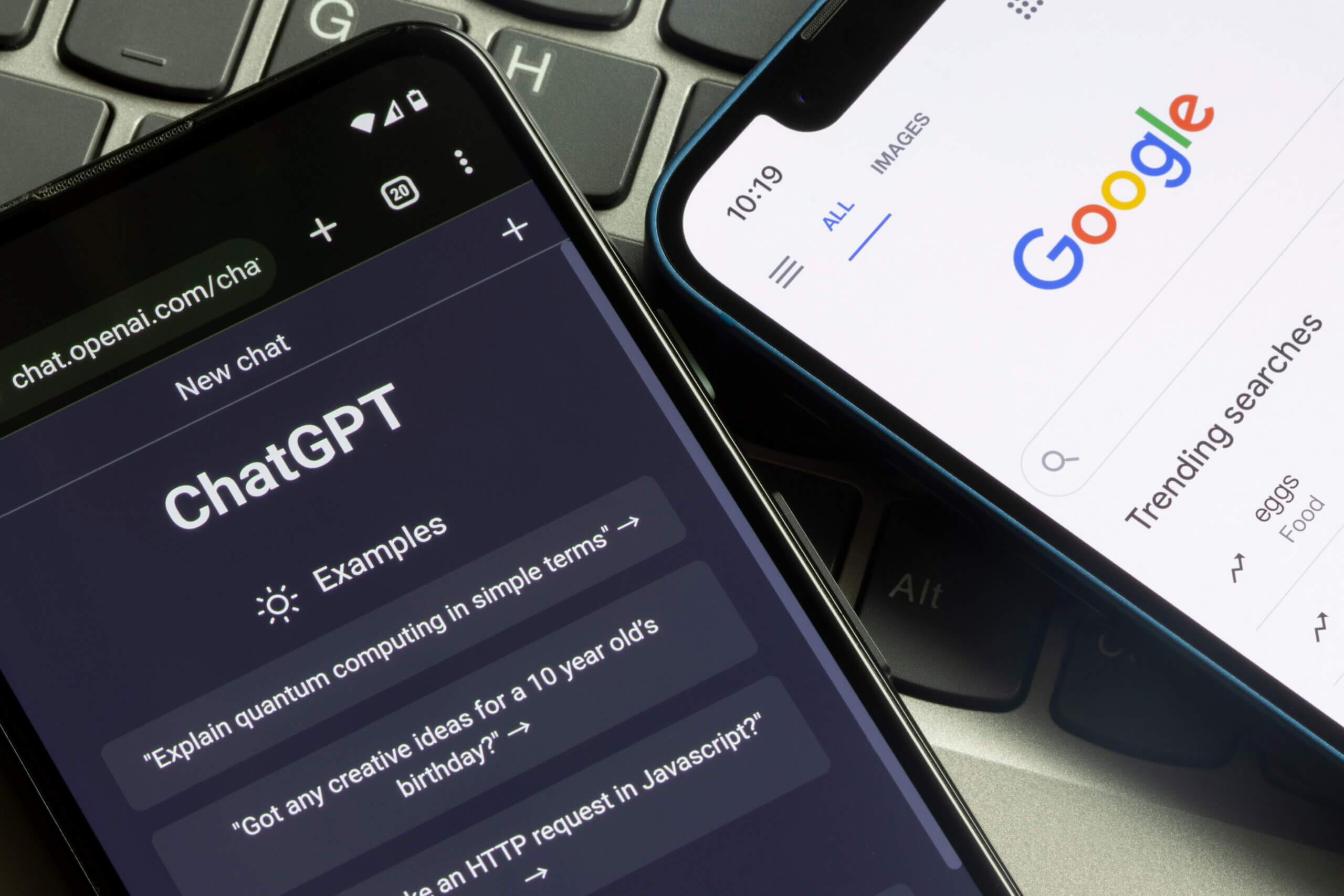 Two smartphones lie on a laptop keyboard. One phone displays the ChatGPT interface on a dark screen, while the other shows a Google search page with the Google logo and search bar on a white background. The devices slightly overlap, highlighting the contrast between the two search and AI tools.