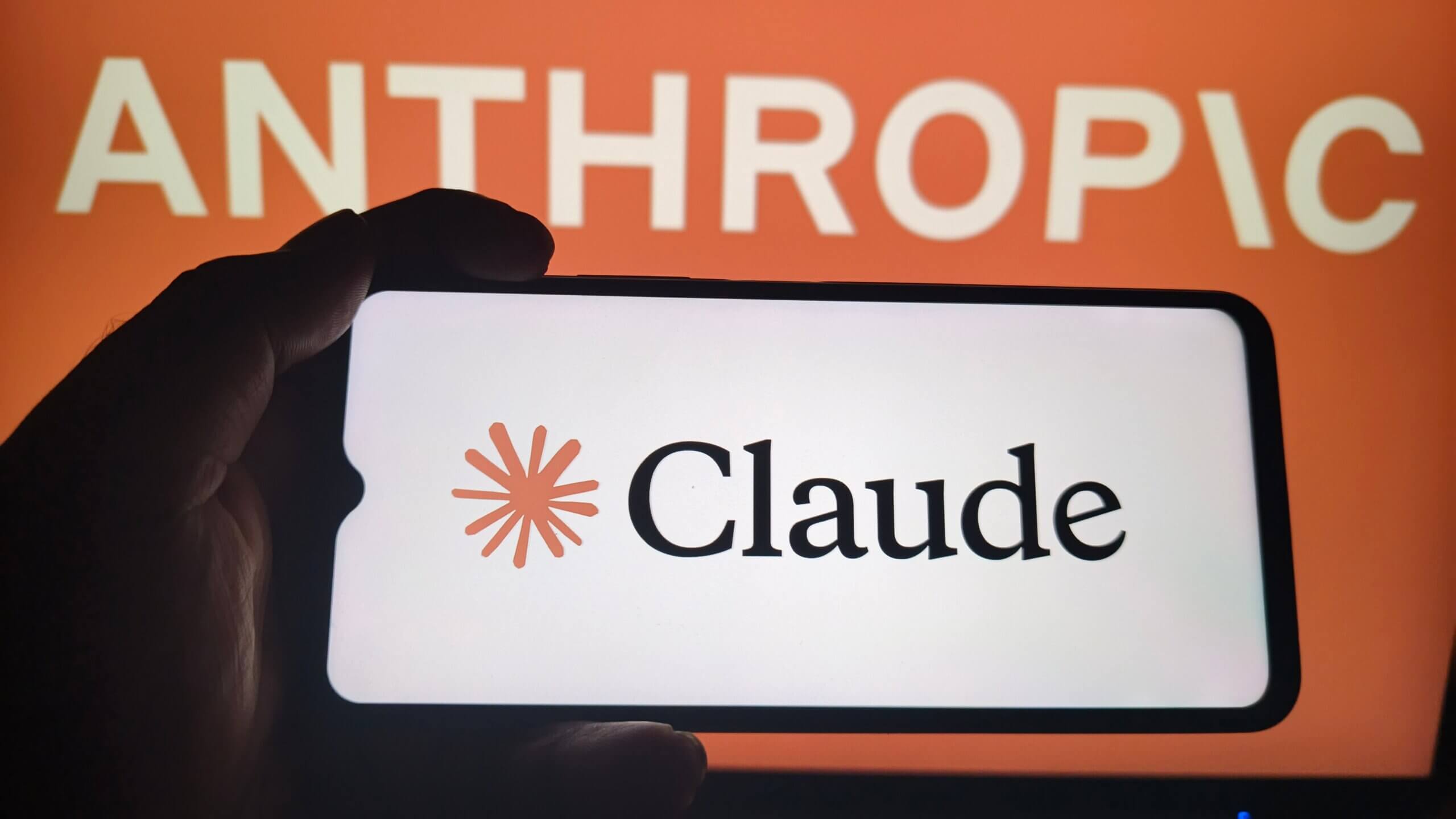 A hand holding a smartphone with the Claude logo on the screen in front of an Anthropic sign