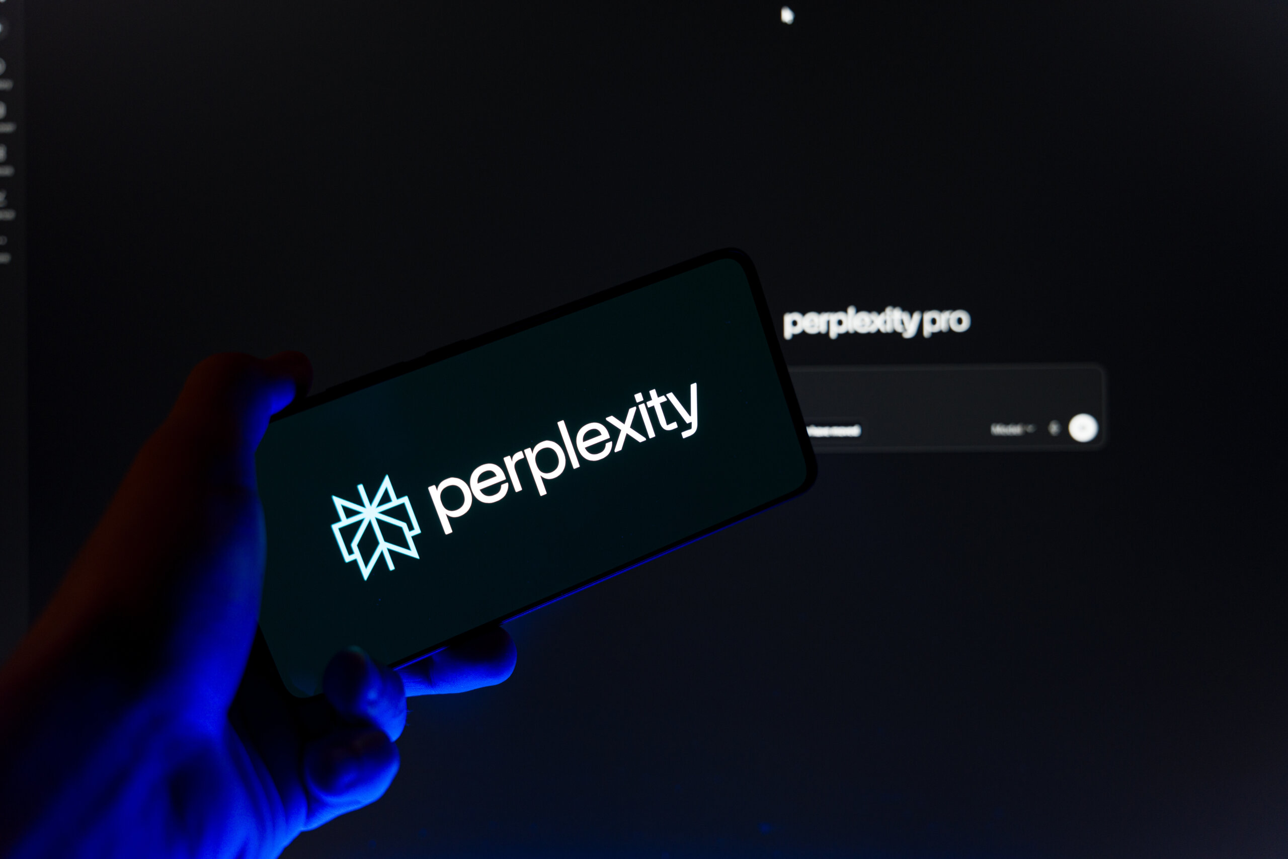 Hand holding smartphone displaying Perplexity AI logo on screen with blurred Perplexity Pro website in background, generative artificial intelligence search engine and research tool concept.