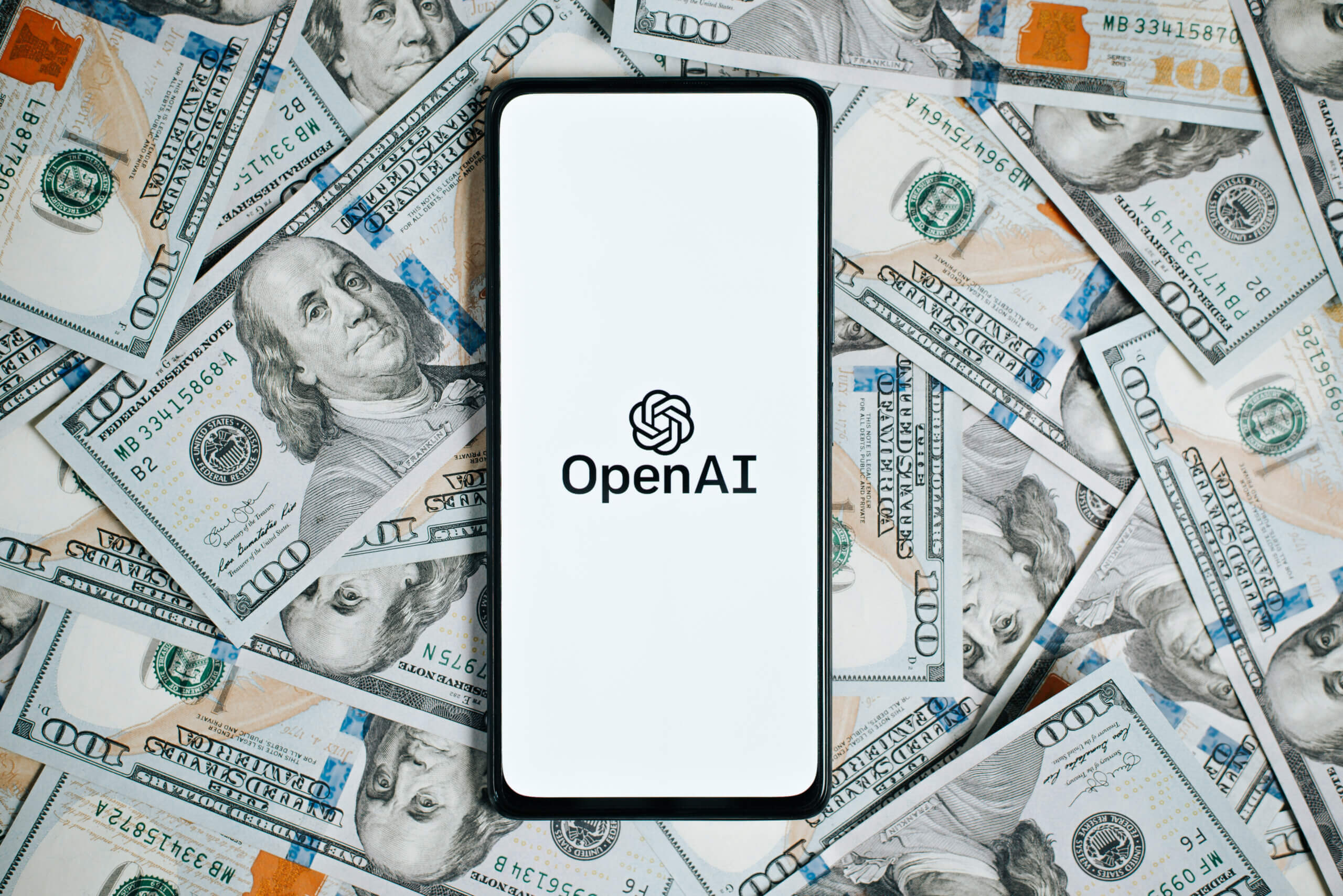 OpenAi company logo on screen of smartphone lying on money background of one hundred dollar bills, top view. Business and technology concept.