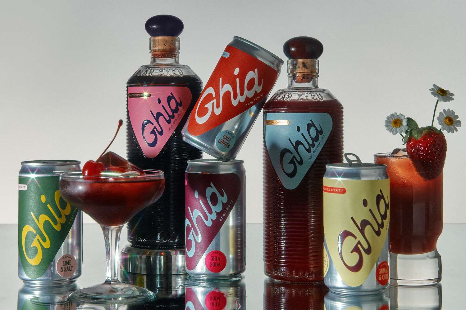 Assortment of Ghia nonalcoholic apéritif bottles and cans styled with two red cocktails, cherries, a strawberry garnish, and small white flowers in a polished studio product shot.