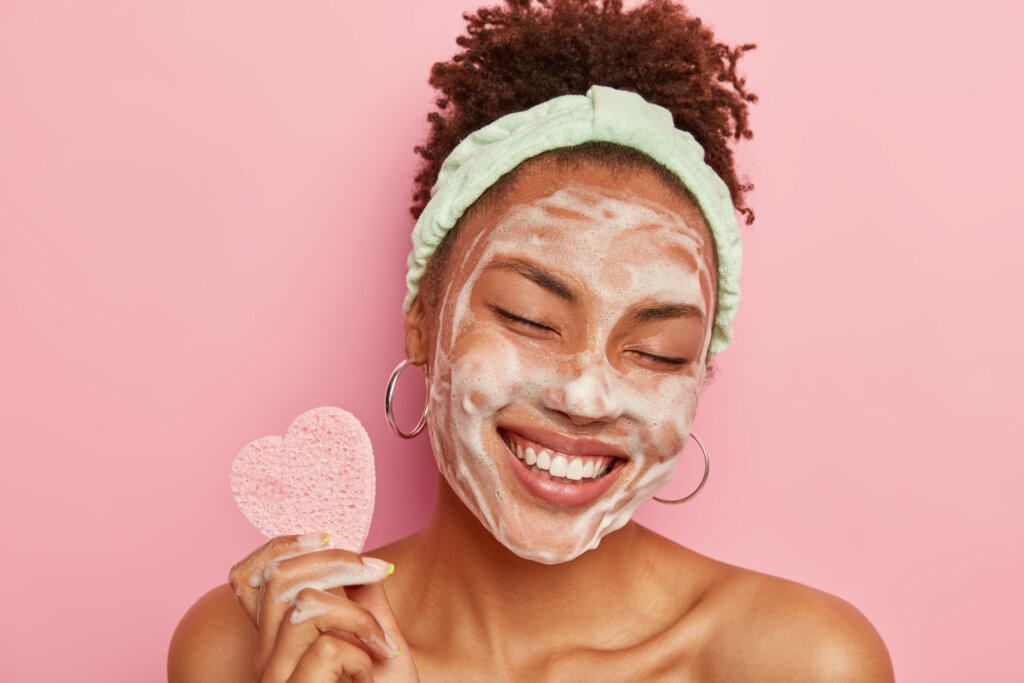 Beautiful optimistic Afro American woman cleanses face with foam, refreshes skin, has well cared complexion, holds heart shaped sponge for beauty procedures, stands bare shoulders with closed eyes